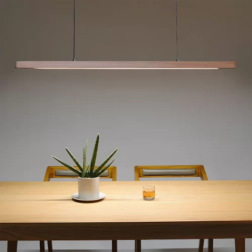 The oval linear ash chandelier casts a warm glow over a wooden table, featuring a potted plant and a cup of tea in the center.